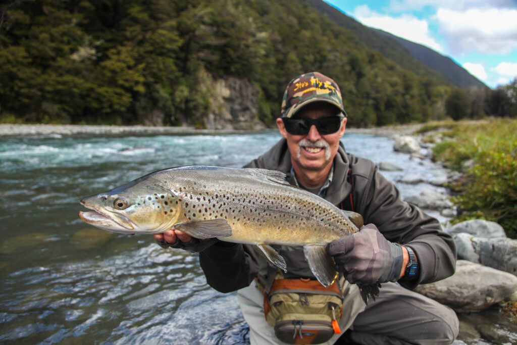 A Man Proudly Holds a Large Brown Trout in a Flowing River, Showcasing His Successful Catch.